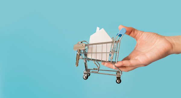 Close up hand holding miniature shopping cart with paper house keys against blue background web